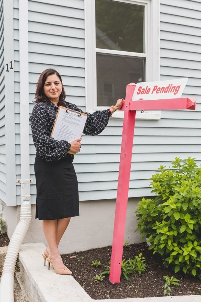 Real Estate Marketing System - Confident female realtor standing next to a Sale Pending sign outside a house, showcasing success.
